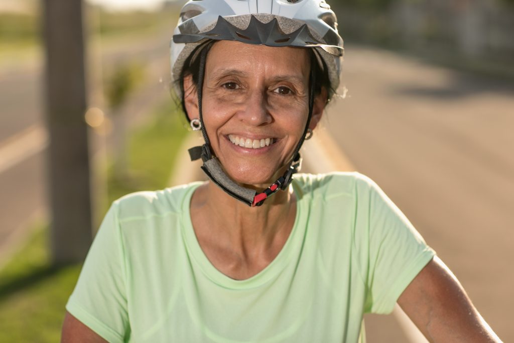 A fotografia mostra Cristina Wayne Nogueira, pesquisadora que atua na área de Farmácia da Universidade Federal de Santa Maria (UFSM). Cristina é uma mulher branca, veste uma blusa na cor verde clara e um capacete. A pesquisadora está com sua bicicleta na ciclovia e sorri.